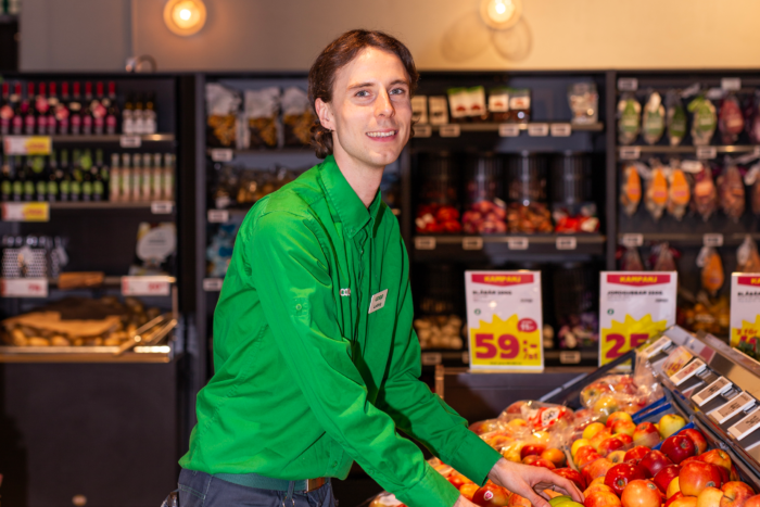 Ung man i Coop-arbetskläder står vid fruktdisken och ordnar äpplen samtidigt som han leende tittar in i kameran.